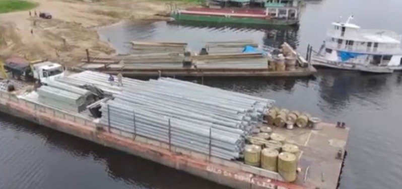 Vista aérea de balsa carregada com postes metálicos e bobinas de cabo em porto fluvial