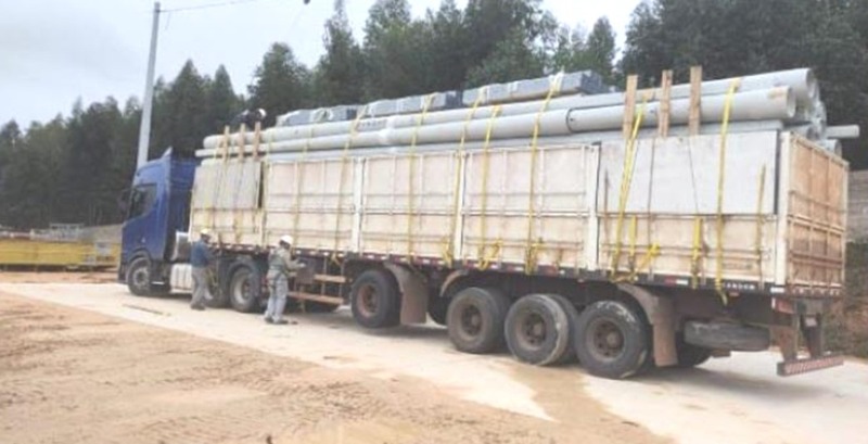 Carreta carregada com postes de concreto e materiais elétricos amarrados, com equipe ao lado